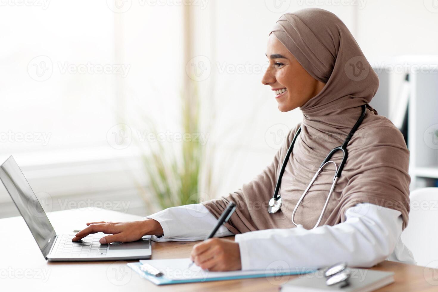 Young female doctor having online training, sitting in her cabinet, using laptop and taking notes, side view, copy space. Attractive muslim woman physician working with computer, clinic interior photo