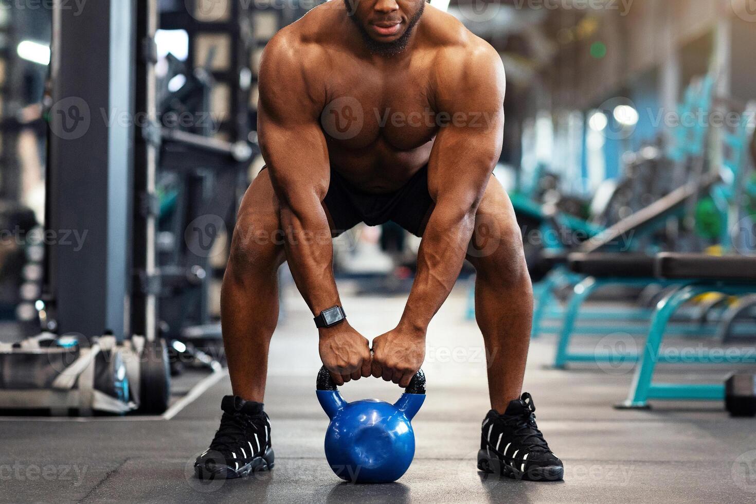 Muscular shirtless african american guy bodybuilder exercising with kettlebell at gym, looking at camera, copy space. Athletic black man making weightlifting or powerlifting, working on body musculs photo