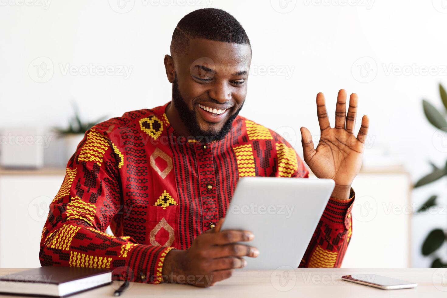 Cheerful black freelancer guy in african shirt making call with digital tablet, sitting at desk in home office, waving hand at camera, greeting somebody, enjoying online communication photo