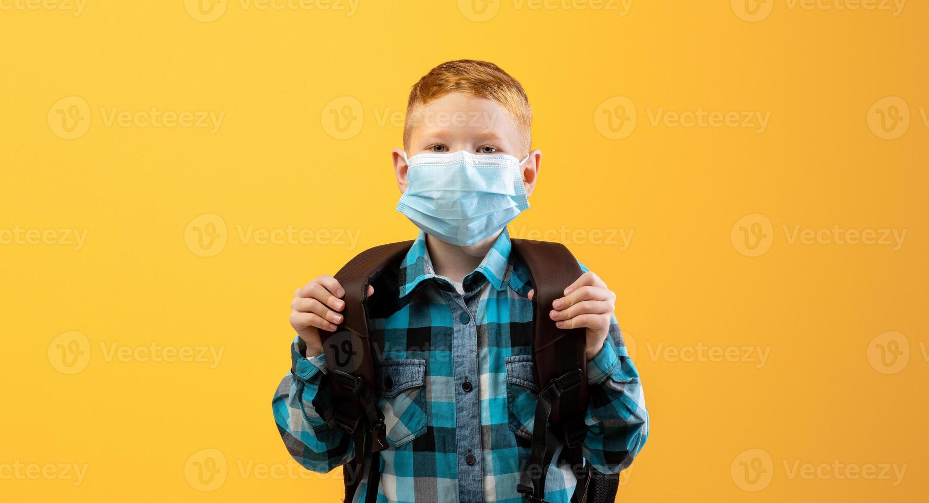 Schoolboy with backpack wearing protective face mask over yellow studio background. Redhead kid schooler using protection against coronavirus while going to school during COVID-19 pandemic photo