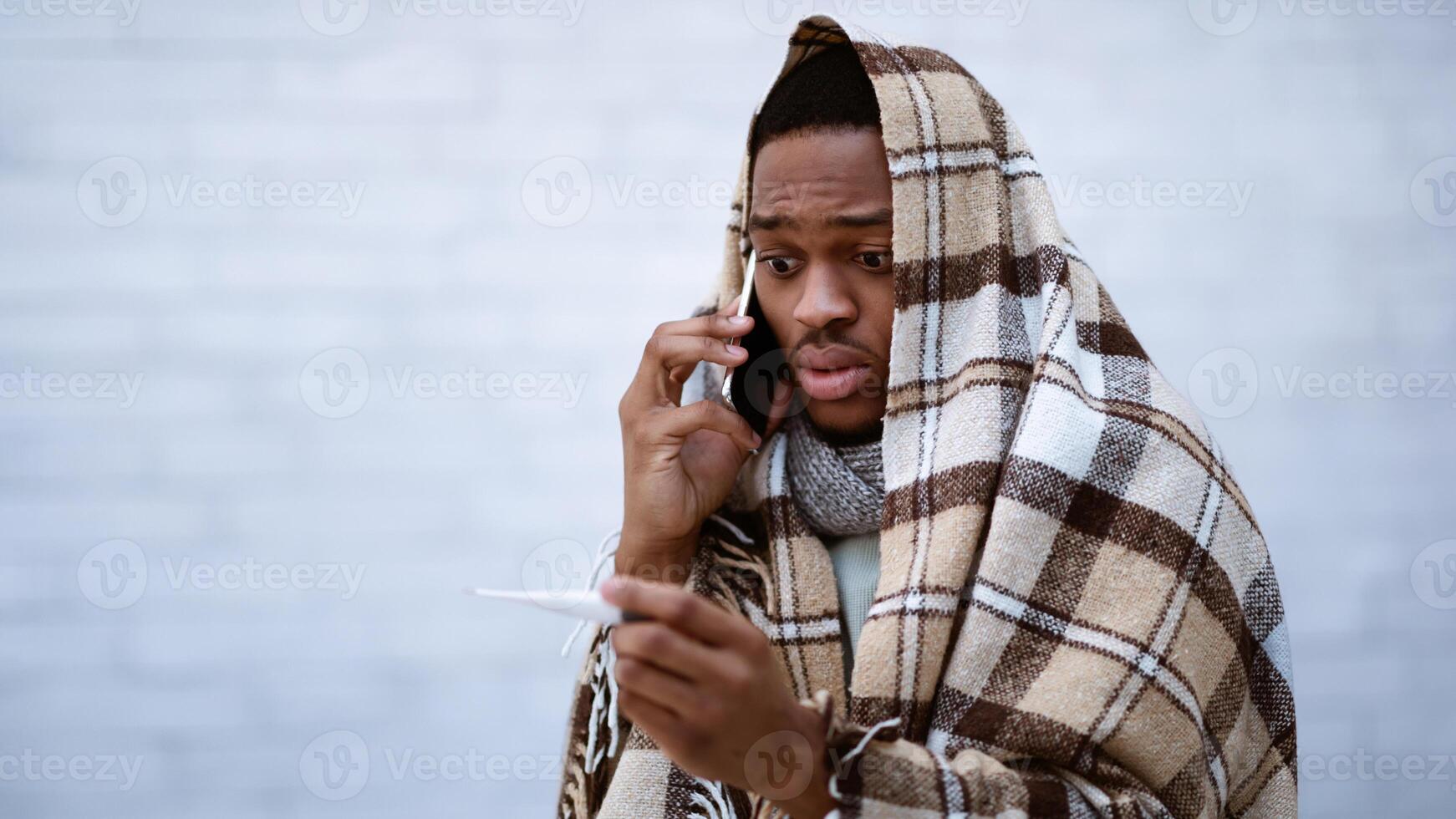 Shocked Sick African American Man Calling Doctor Having Fever Standing Wrapped In Blanket Over Gray Wall Background. Covid-19 Corona Virus, Cold And Influenza Symptom. Selective Focus photo