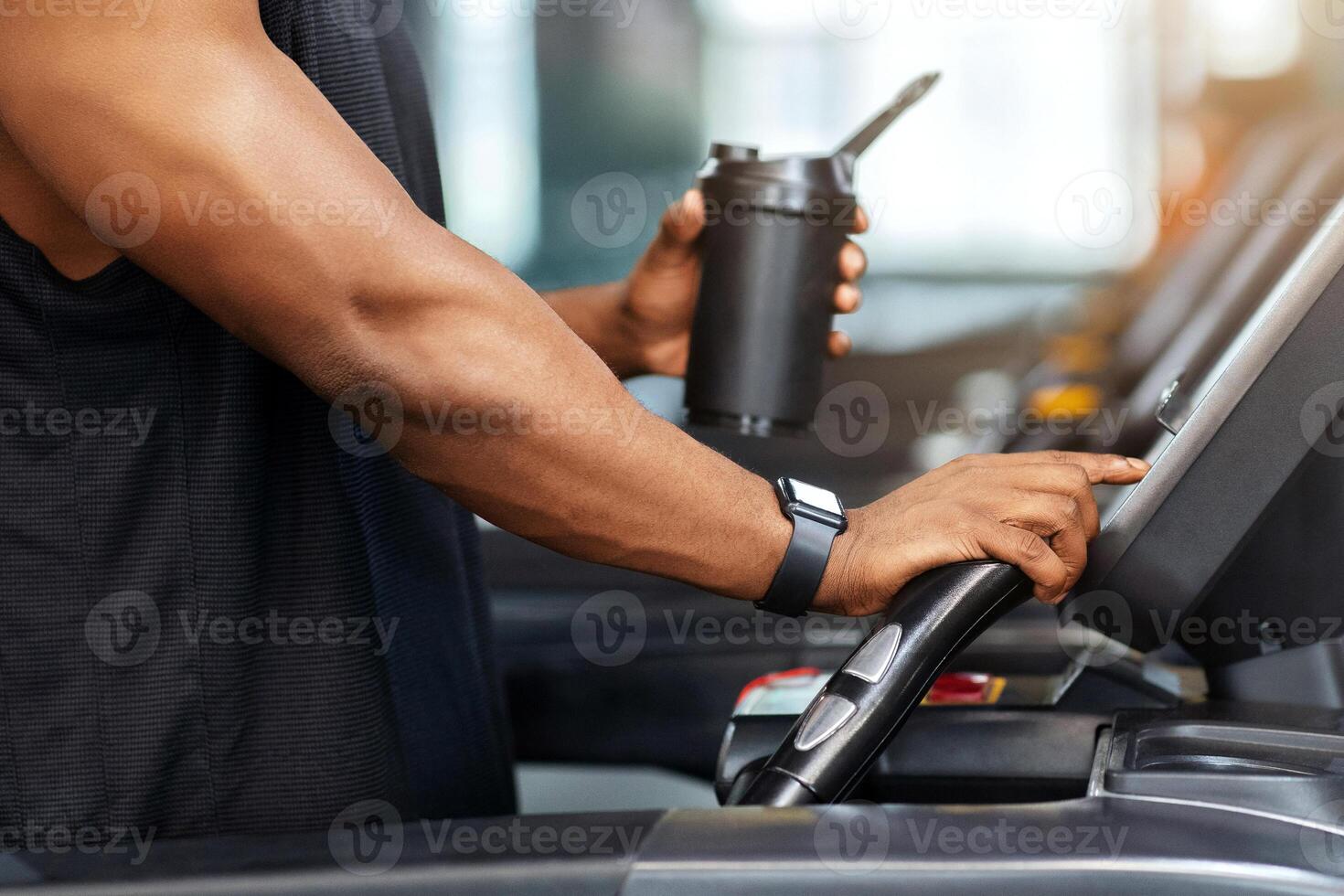 Cropped of muscular african american man holding sports drink, standing on treadmill, drinking protein before running, copy space. Unrecognizable black guy bodybuilder jogging at gym photo