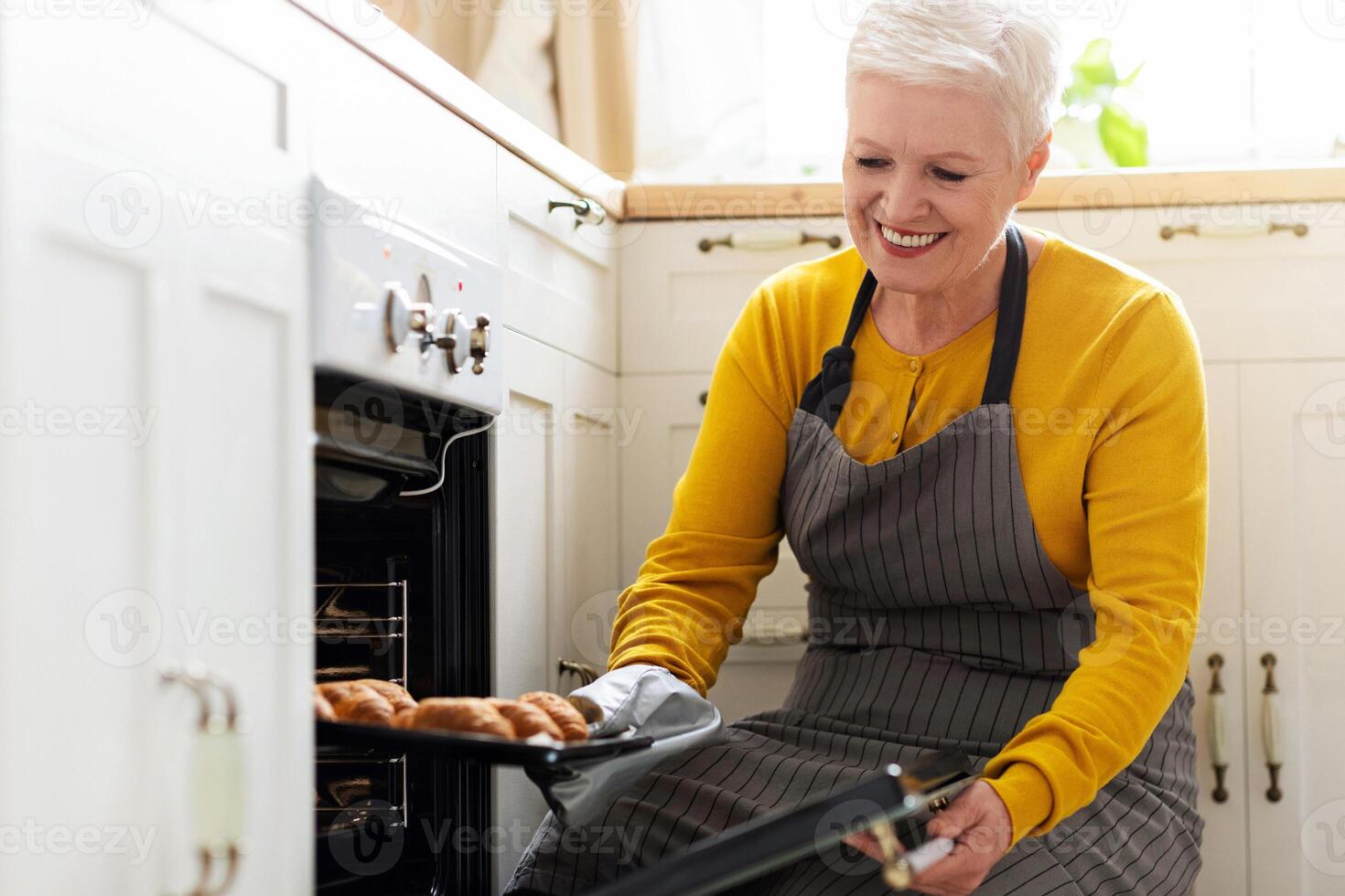 Joyful senior lady in apron opening oven, checking her delicious croissants, cozy kitchen interior, copy space. Smiling elderly woman baking pastry at home, taking cookies out of oven photo