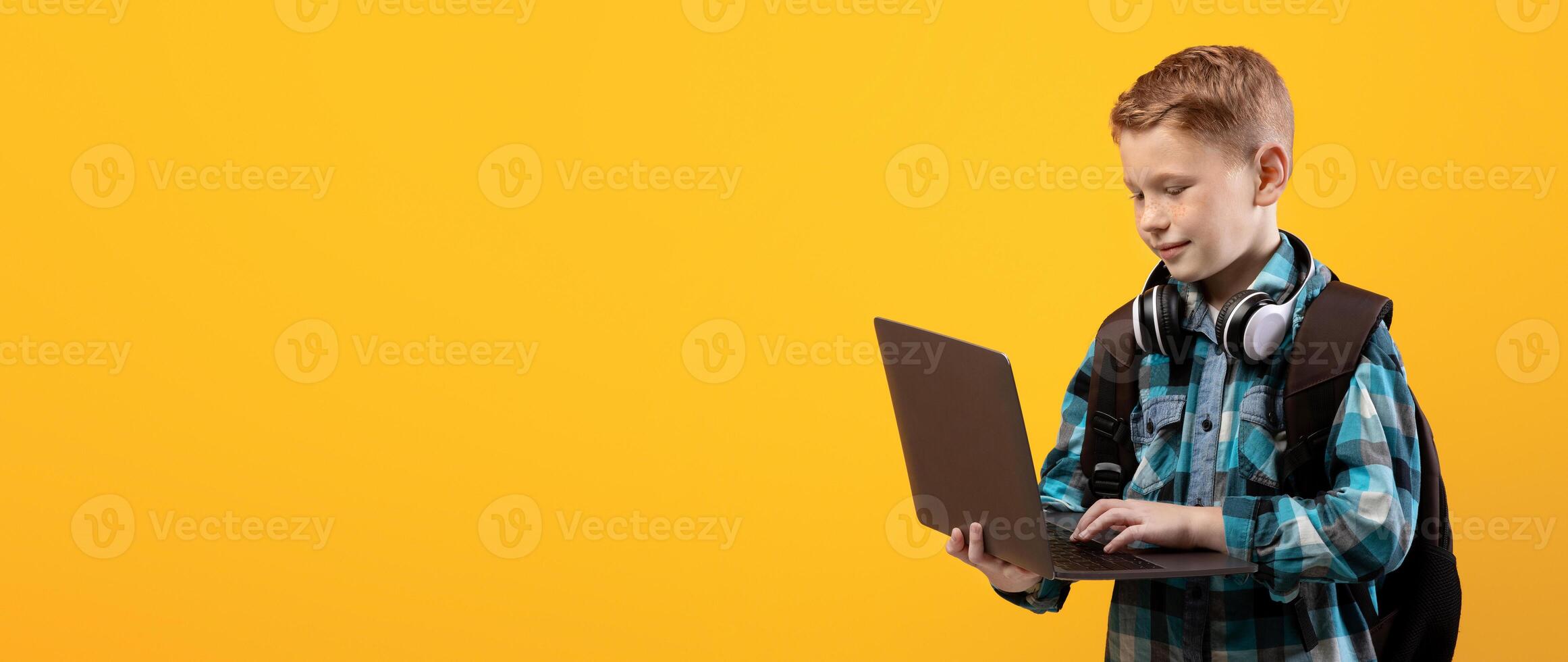 Technologies in school education. Smart schoolboy with backpack and headset holding laptop over yellow studio background, copy space. Cute kid using laptop for studying, doing homework, websurfing photo