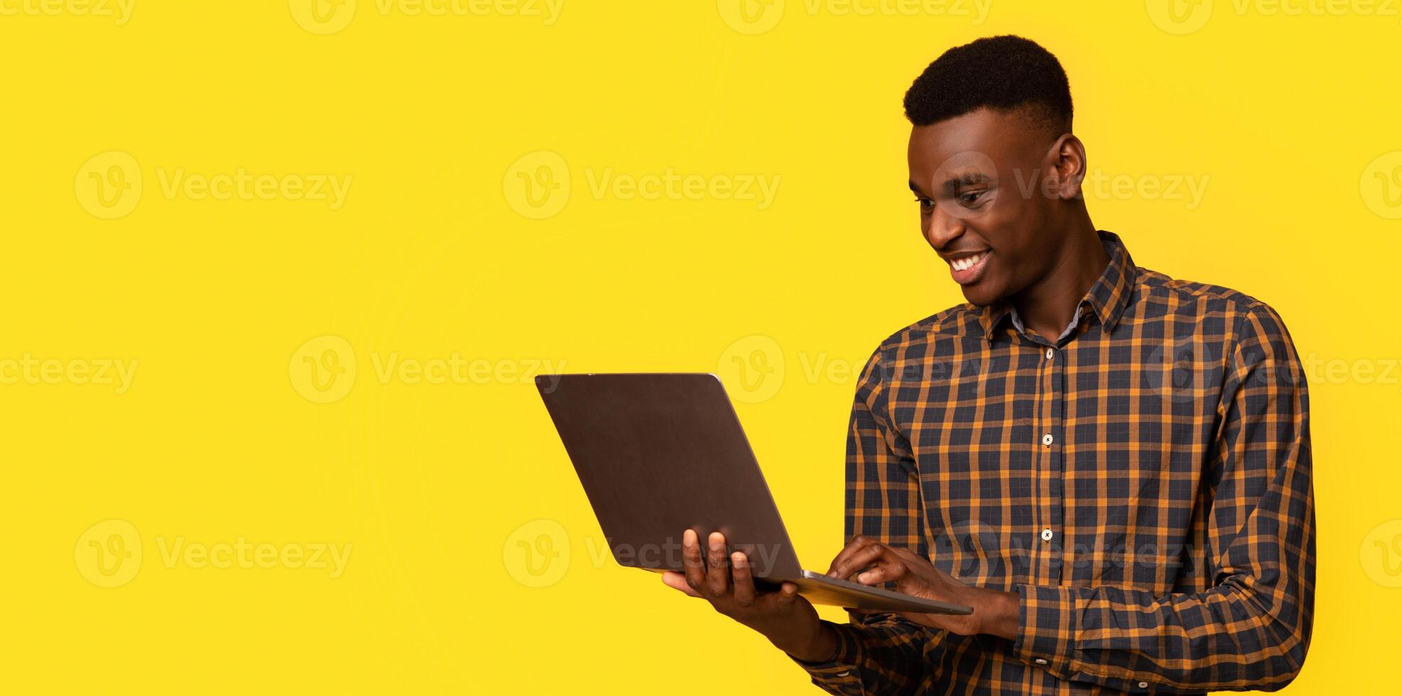 Freelance Work. Portrait Of Smiling African American Guy Using Laptop While Standing Isolated Over Yellow Background, Black Millennial Guy Working Online, Enjoying Remote Job Opportunities, Copy Space photo