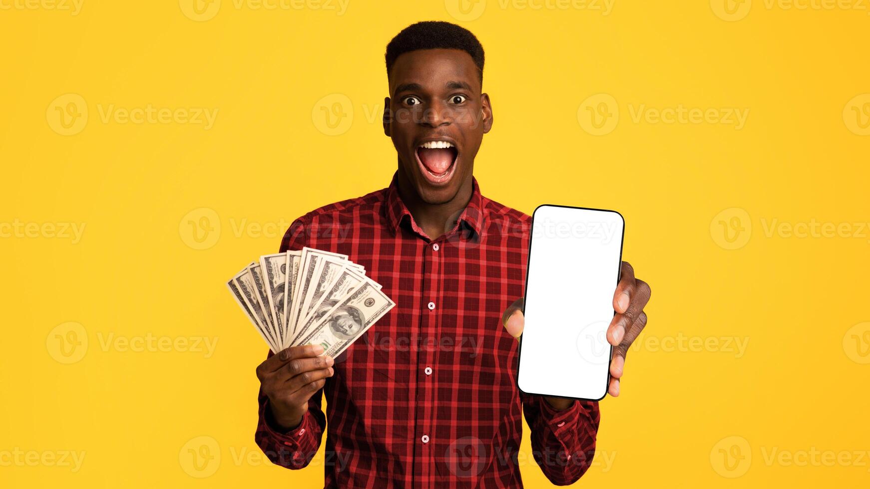 Surprised black guy holding smartphone with blank screen and dollar cash money in hand, excited african man emotionally recting to big win, posing isolated on yellow background, mockup image photo