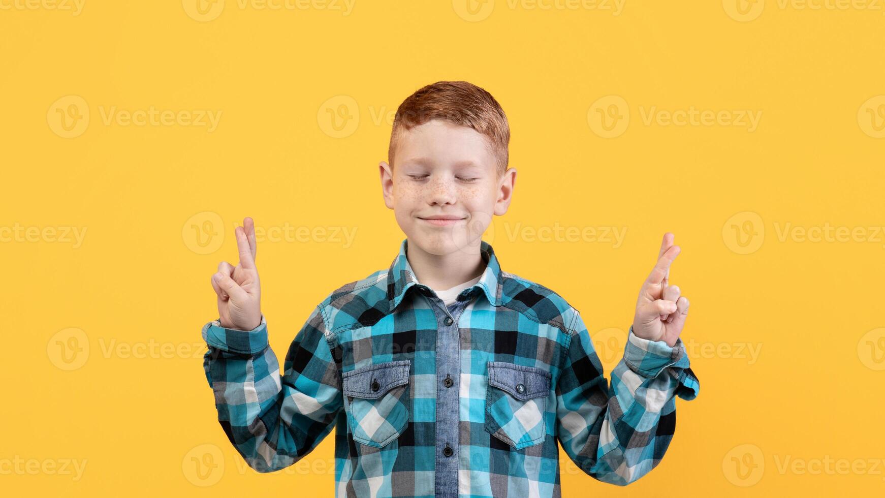 Positive kid with eyes closed showing crossed fingers on yellow studio background, copy space. Little ginger boy in cute shirt hoping or praying for best, holding fingers crossed, dreaming photo