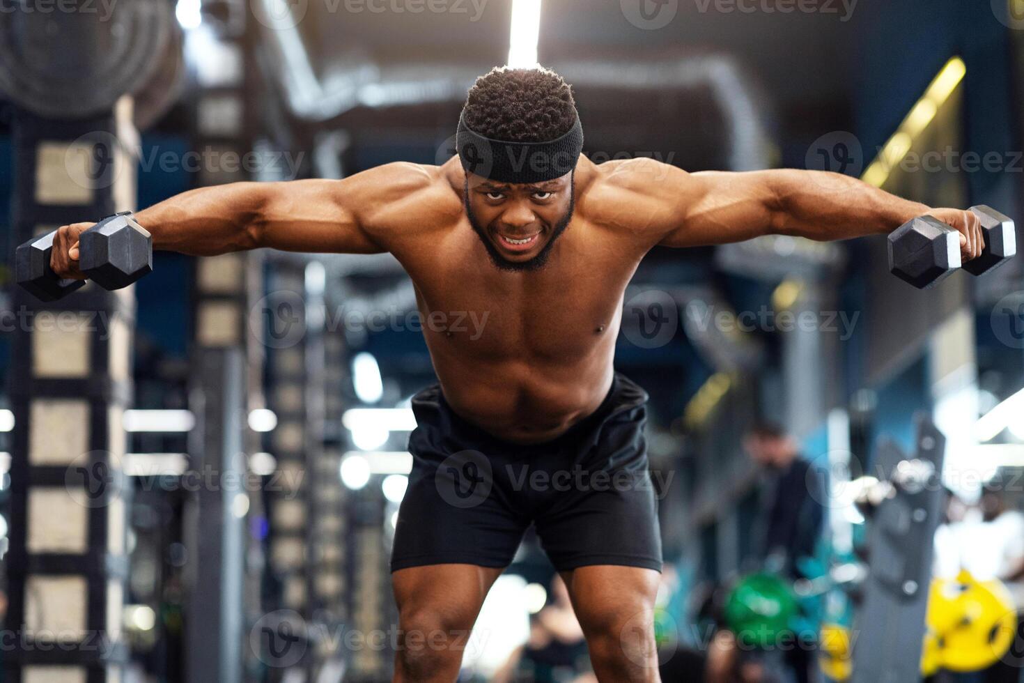 Strong shirtless black guy bodybuilder making workout with dumbbells at gym. Emotional african american athletic man lifting barbells up, working on arms and back muscles, copy space photo