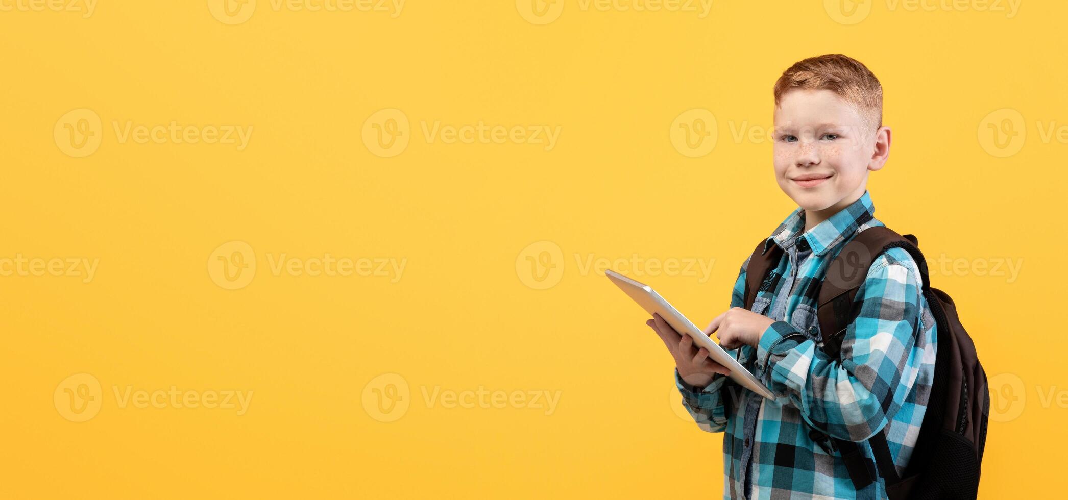 Cheerful schoolboy using digital tablet for education, doing homework on yellow studio background, copy space. Smiling redhead boy with backpack holding pad, websurfing or using new application photo