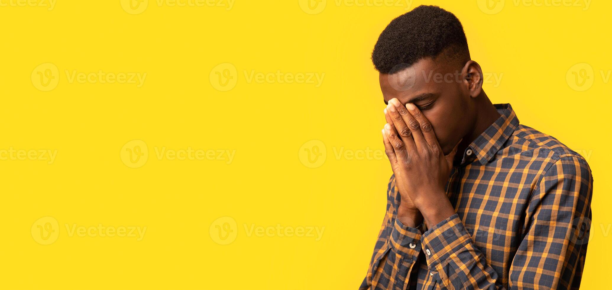 Upset black guy covering face with two hands in despair, feeling stressed and depressed, lonely sad millennial african american man standing isolated over yellow background, closeup shot, copy space photo