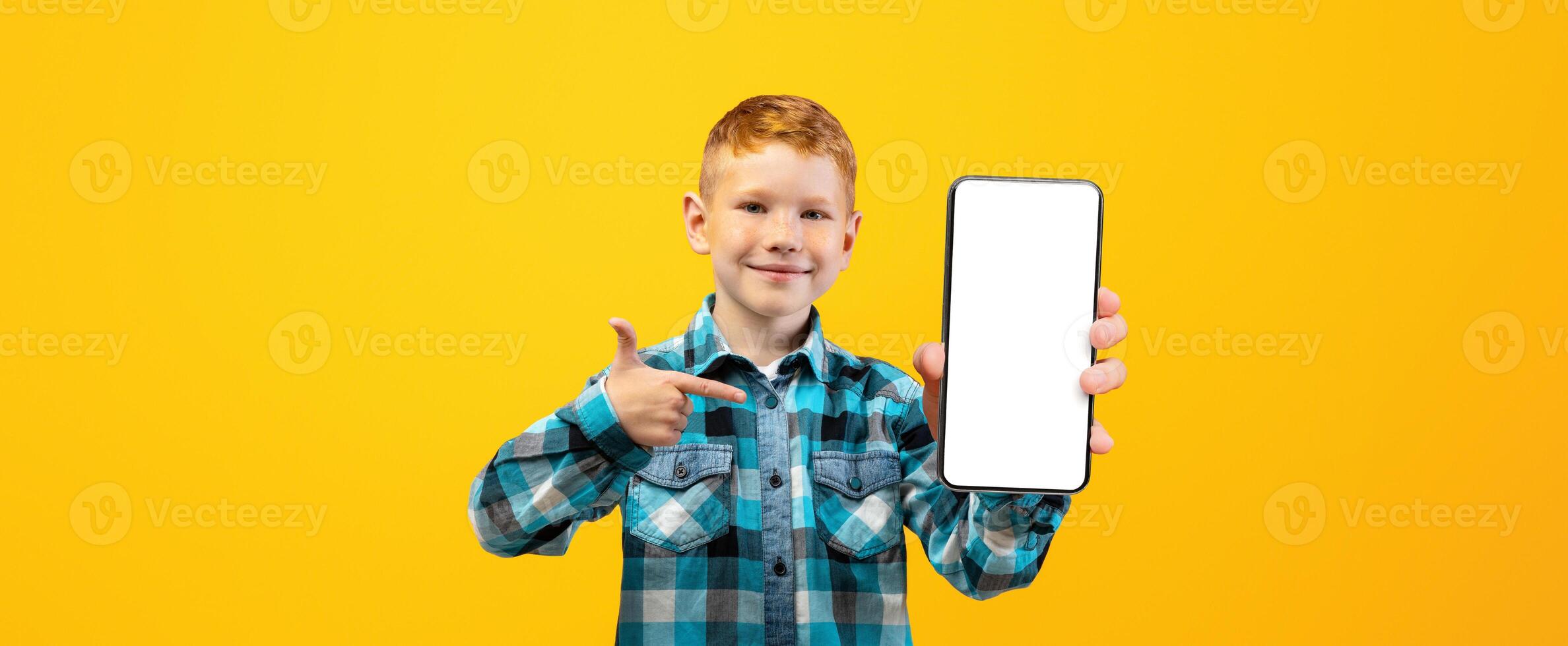 Smiling ginger boy in casual shirt pointing at modern mobile phone with empty screen over yellow studio background, mockup. Happy schoolboy with smartphone for education or entertainment, copy space photo