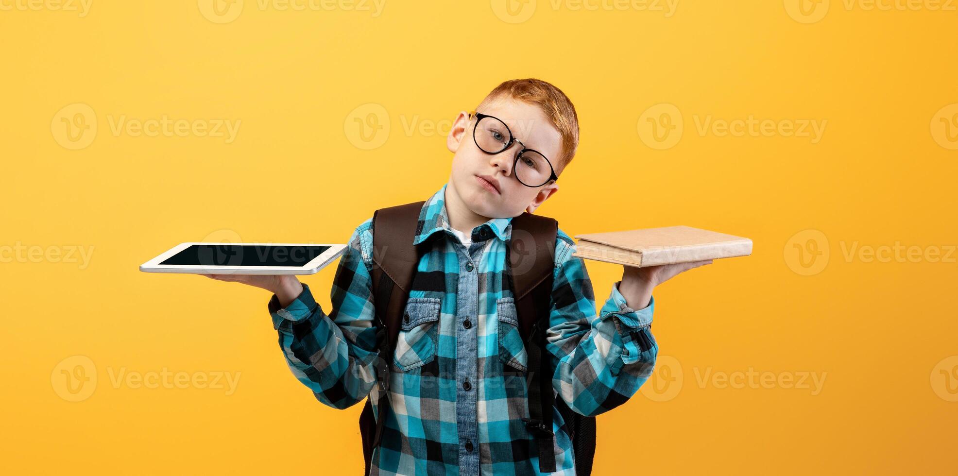 Confused kid schooler with backpack and glasses holding digital tablet and book, choosing between studying and playing, yellow studio background, copy space. Paper book vs digital tablet photo