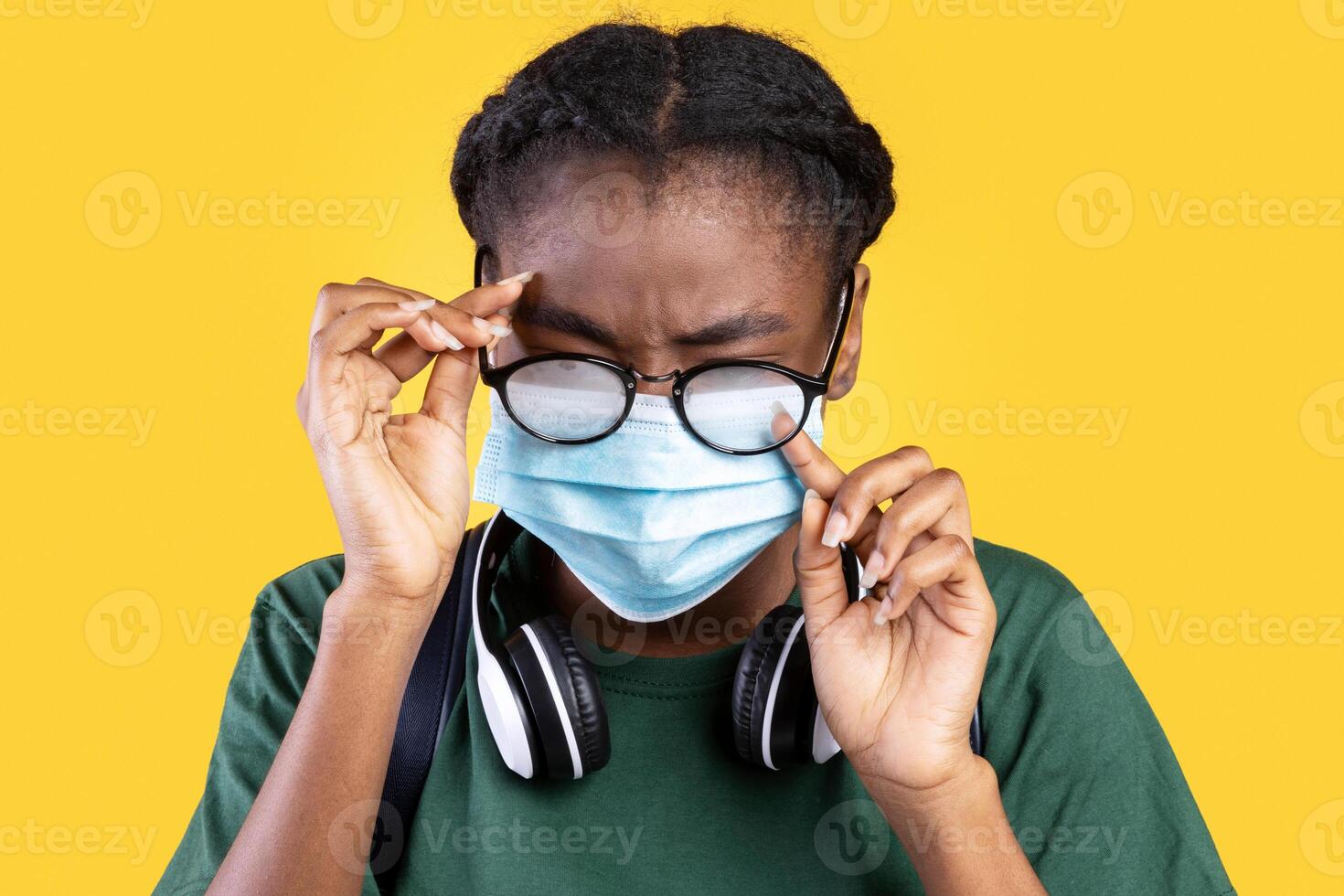 Don't Touch Your Eyes. African Female Student Scratching Eye Wearing Medical Face Mask Standing On Yellow Background In Studio. Coronavirus Prevention, Covid-19 Protection During Pandemic Outbreak photo