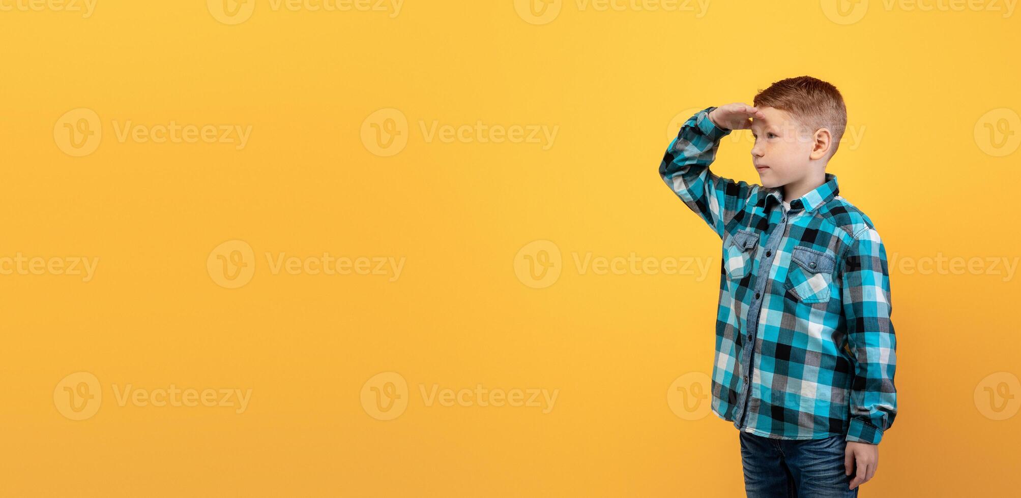 Cute ginger kid in flannel holding palm next to forehead, looking at copy space, panorama, yellow studio background. Curious red-haired boy watching exciting advertisement for kids photo