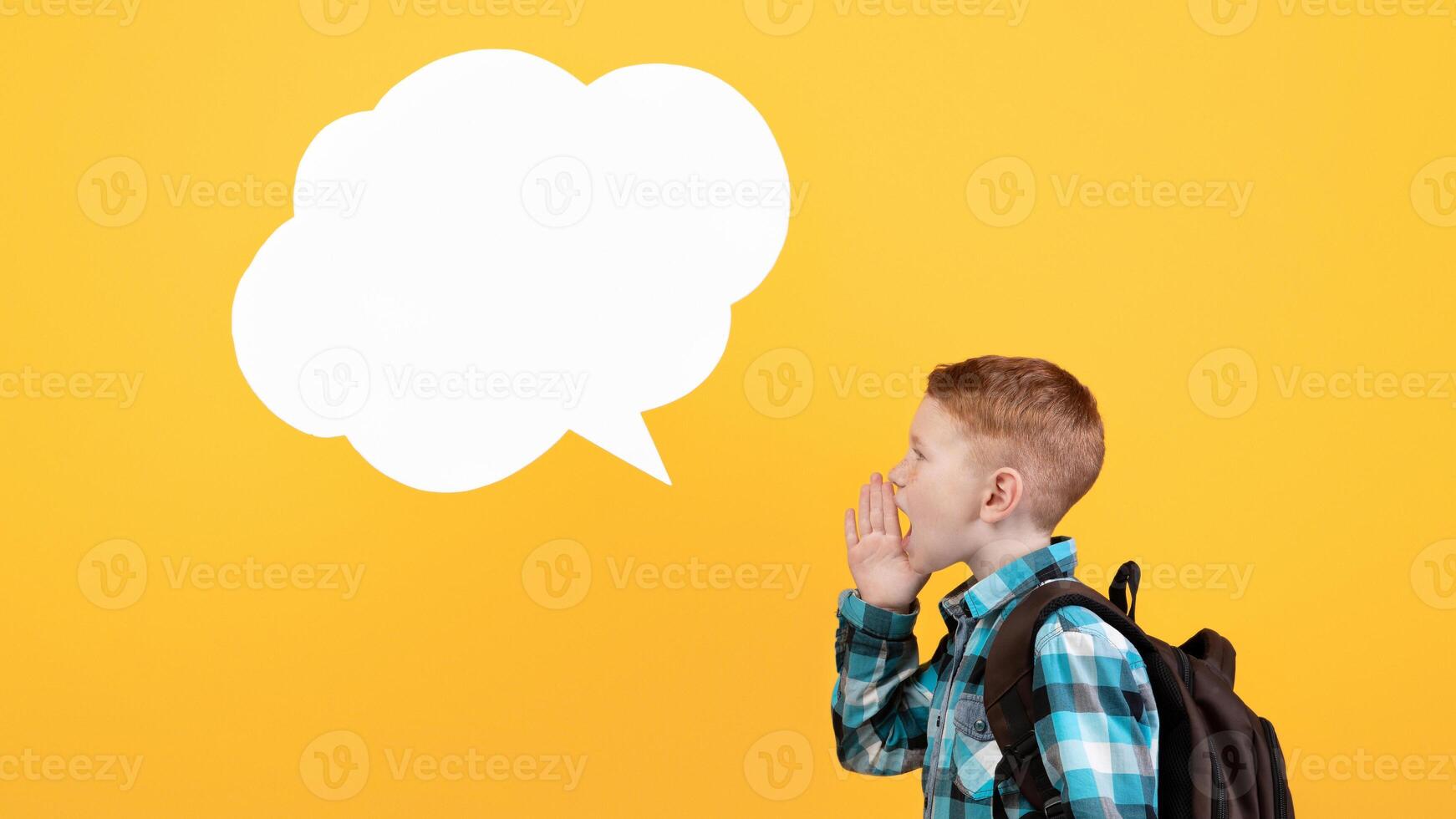 Side view of ginger schoolboy with backpack saying something loud and empty communication cloud over yellow studio background, copy space. Boy with schoolbag looking for attention photo