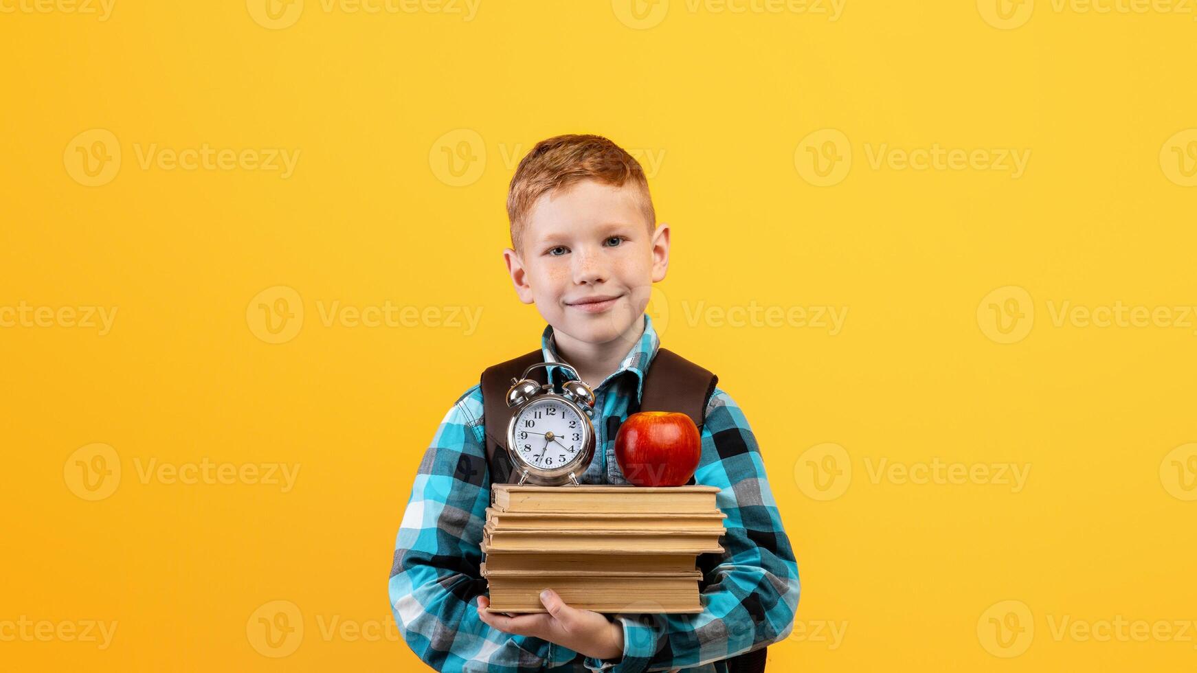 Cheerful ginger kid schooler with backpack holding heap of books, red apple and watch on yellow studio background, copy space. Schedule, timing for children schoolers concept photo