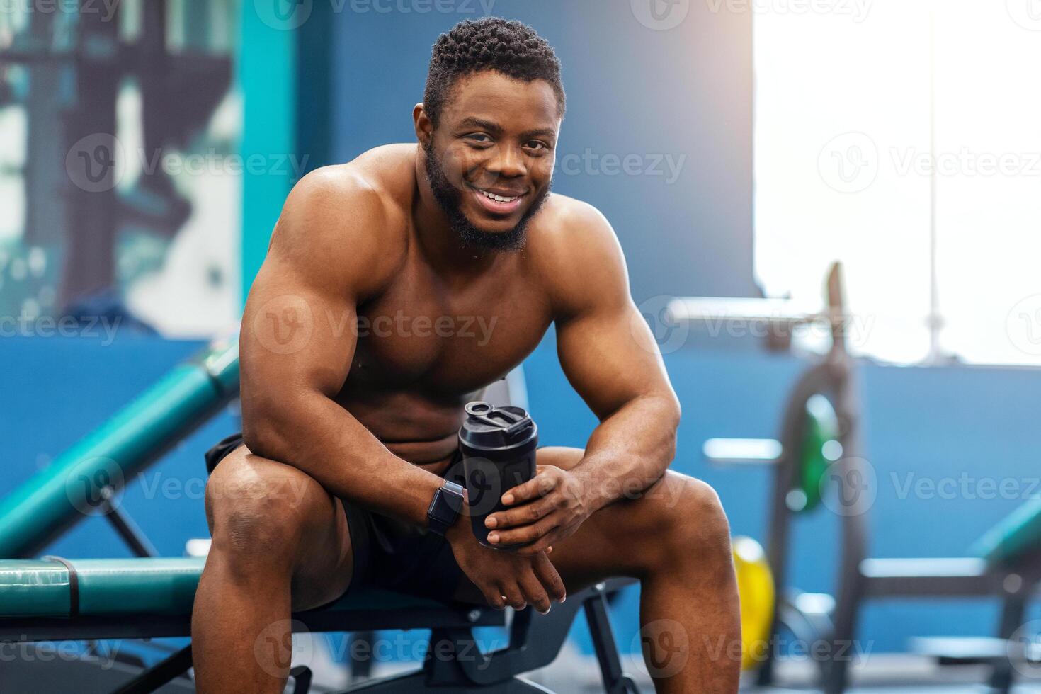 Smiling african american shirtless bodybuilder drinking protein or water while having break after workout at modern gym. Happy black guy sportsman resting after exercising, copy space photo
