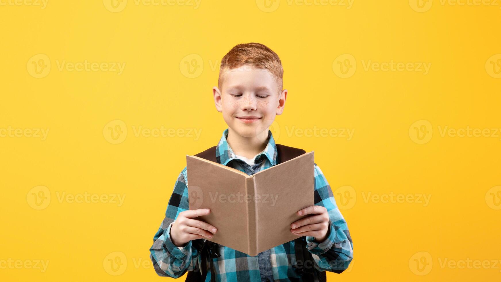 Joyful redhead schoolboy in shirt reading book, yellow studio background. Happy kid with backpack enjoying exciting book, love studying, copy space. School educational system concept photo