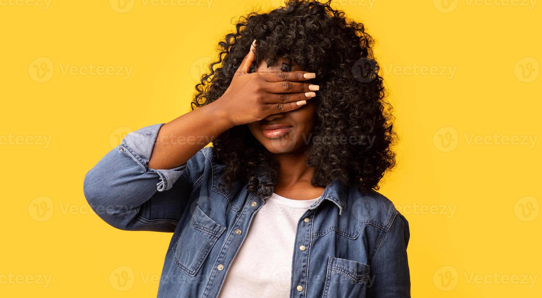 Curly black woman covering eyes with one hand, feeling scared or anxious, wondering or blindly waiting for a surprise against yellow studio background, panorama with copy space photo