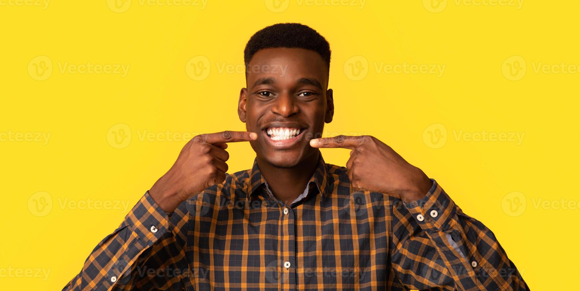 Perfect Smile. Happy Black Millennial Guy Pointing At His White Teeth With Fingers, Recommending Dental Alignment While Standing Isolated Over Yellow Background, Closeup Shot With Copy Space photo