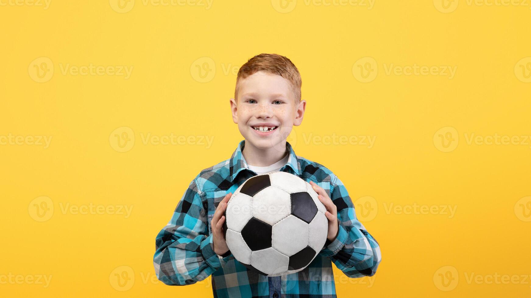 Young football player. Cheerful boy posing with soccer ball on yellow studio background, copy space. Happy kid showing his soccer ball to camera, kids and teens sport lifestyle concept photo