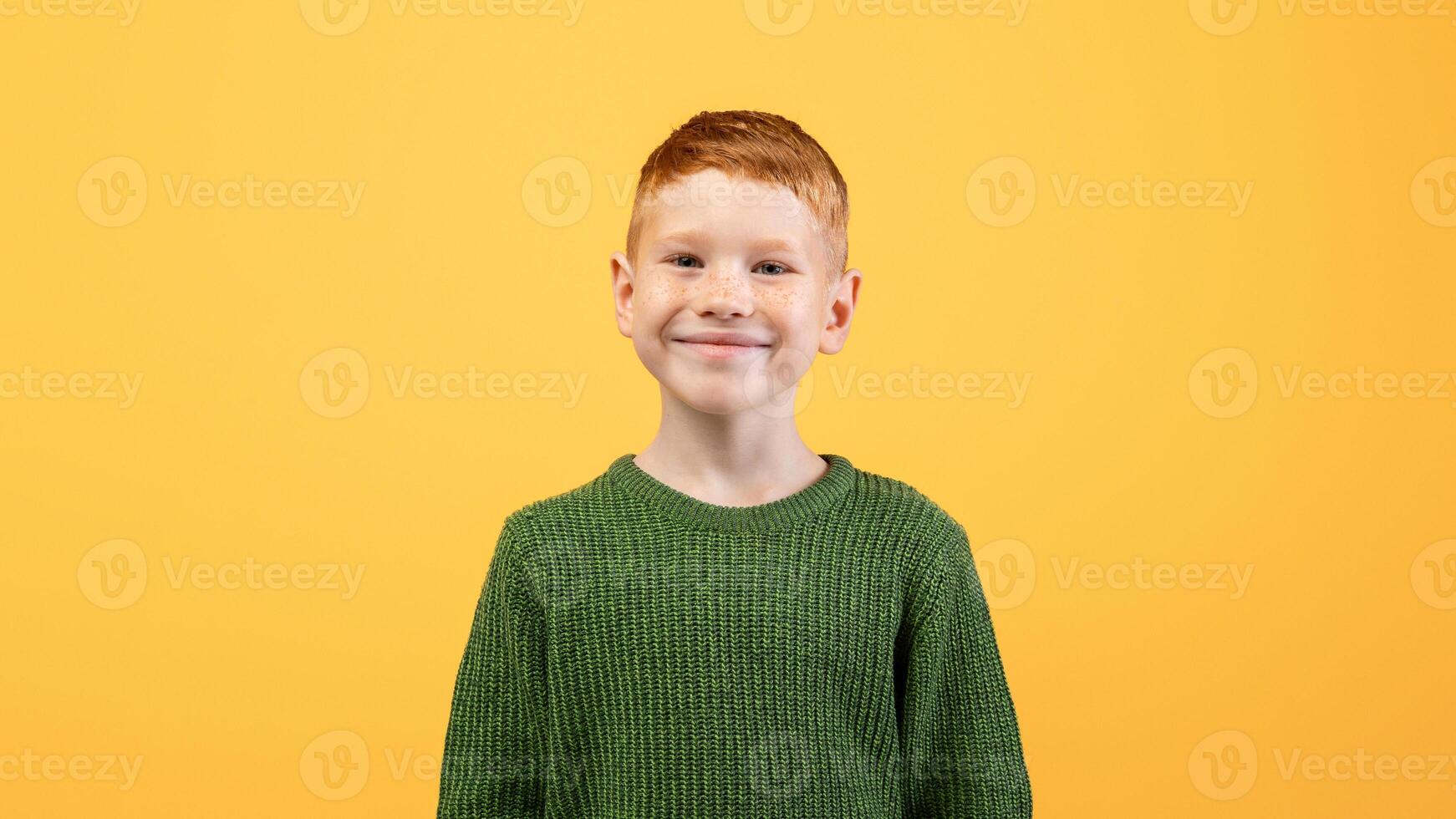 Portrait of happy cute ginger boy in knitted green sweater posing on yellow studio background, copy space. Redhead kid in casual outfit smiling at camera, having good mood. Childhood concept photo