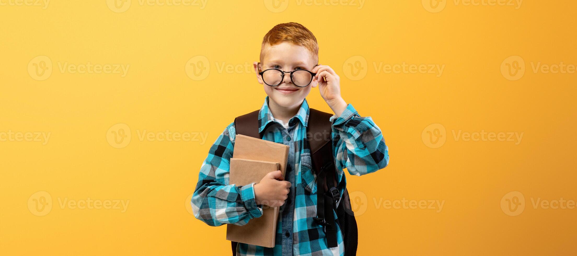 Happy schooler with backpack pulling down glasses, holding heap of books, yellow studio background, copy space. Happy boy going to school, holding books, enjoying school days. Kids education concept photo