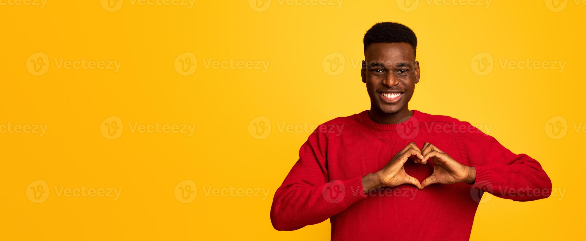Kind black guy shaping hands like heart, making love gesture near chest, friendly african american man expressing kindness, smiling at camera, standing over yellow studio background , free space photo