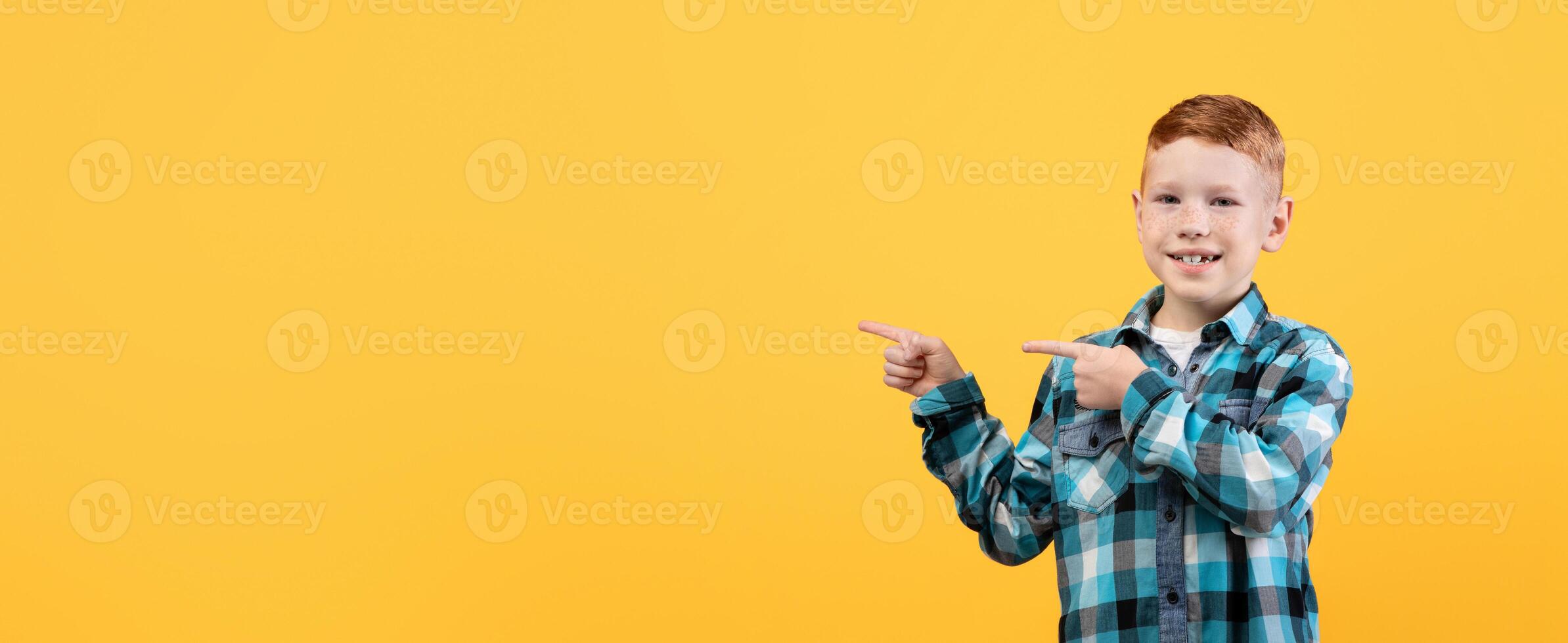 Cute ginger boy in casual outfit pointing with both hands at empty space for text or advertising over yellow studio background. Smiling kid showing exciting advertisement, aiming at free space photo