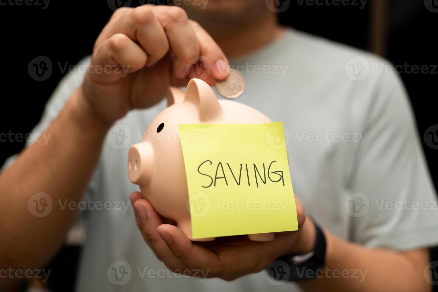 Man Depositing Coin into Piggy Bank with Saving Note. A man's hand is shown actively depositing a coin into a pink piggy bank. financial planning, making deposits, and setting financial goals. photo