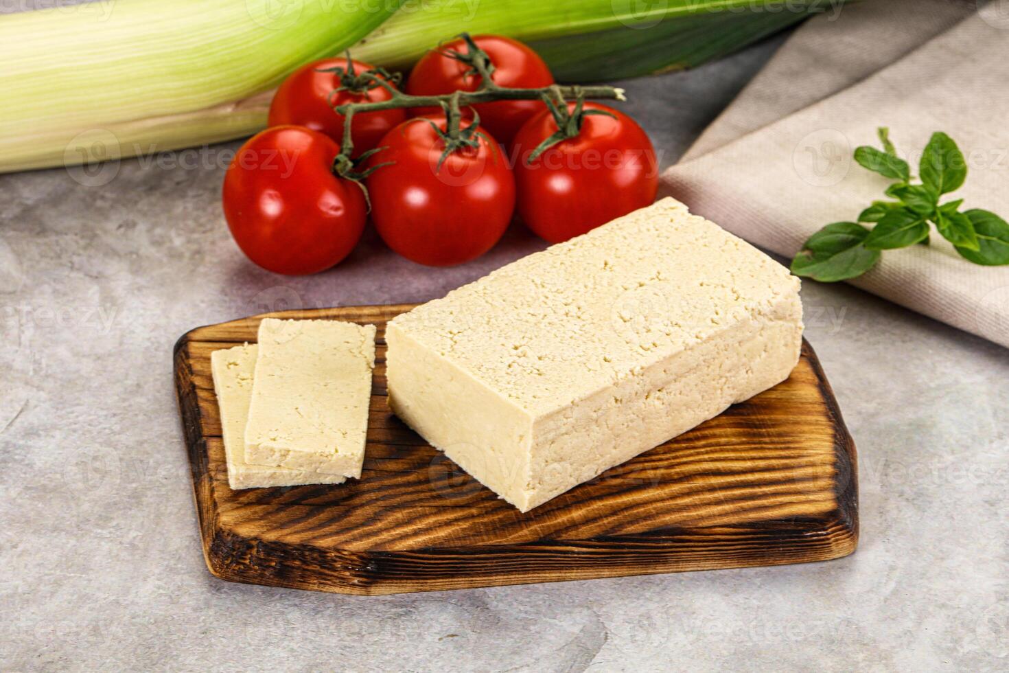 A piece of tofu on a cutting board next to a tomato and a bunch of vegetables photo
