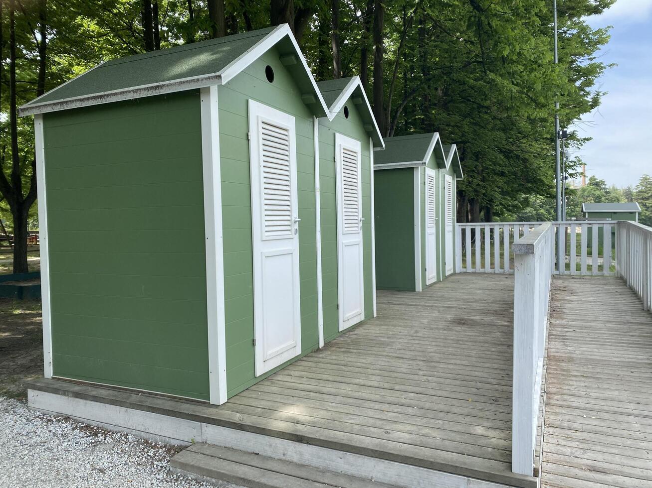 Row of quaint green beach changing huts on a wooden boardwalk beside lush green trees. photo