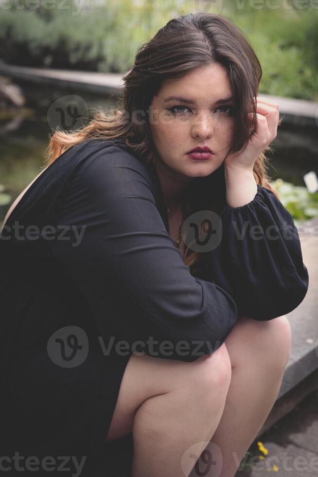 Subject is sitting by a serene pond in a natural setting during a bright day highlighting emotional expression and tranquility photo