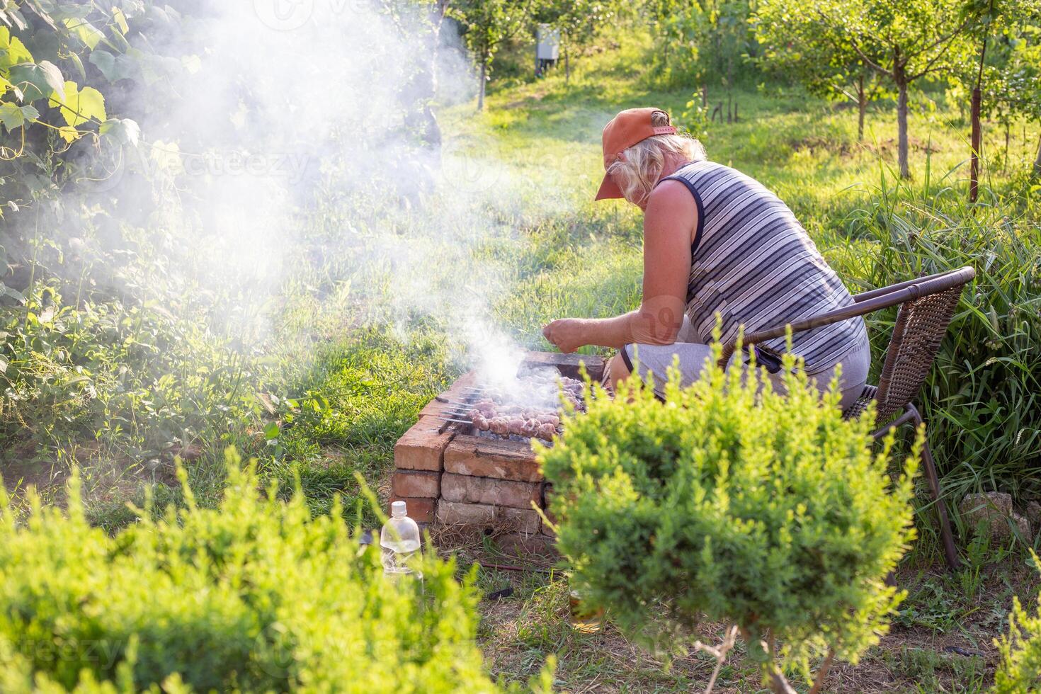 man turns skewers of fresh pork meat on a brick barbecue in the yard of a house. Outdoor picnic with grilled shashlik photo