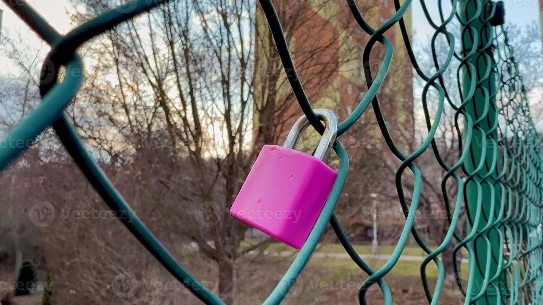 A bright and vibrant pink padlock is securely attached to a sturdy chain link fence, set against a serene backdrop photo