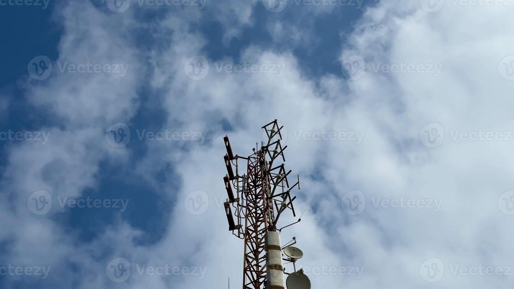 The image shows a telecom tower against a bright blue sky with fluffy clouds, highlighting its antennas photo