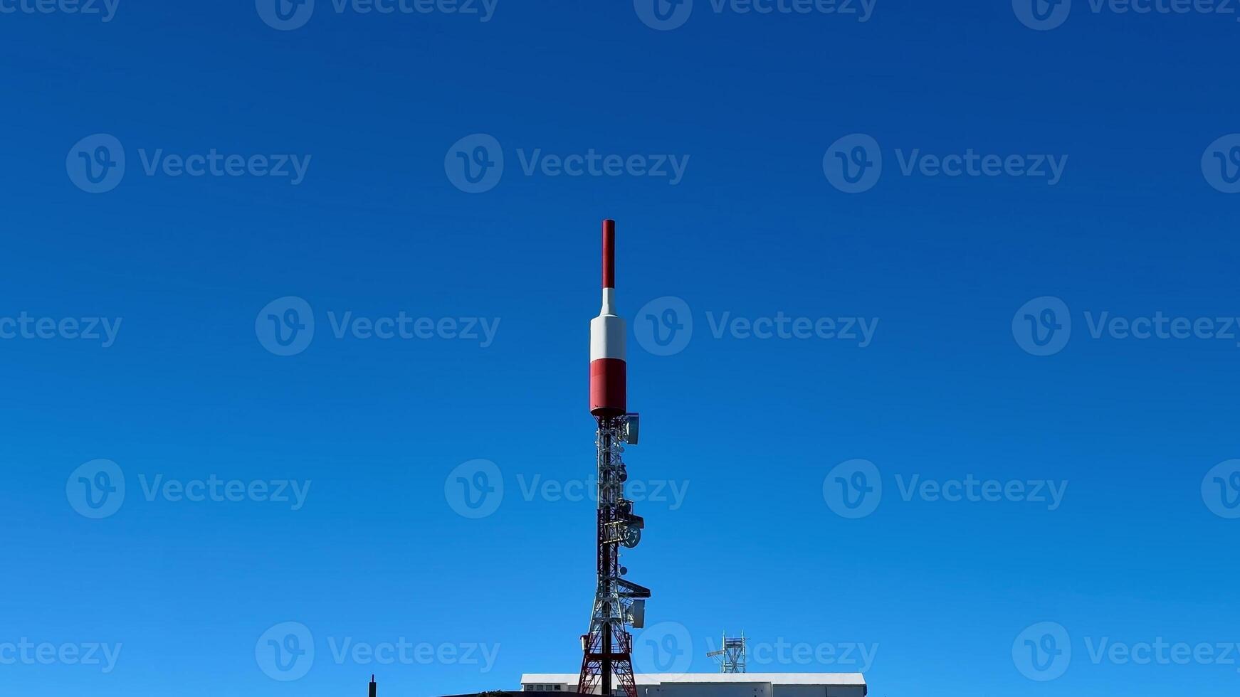 A striking red and white telecom tower prominently stands out on a lush green hillside beneath a bright clear blue sky photo