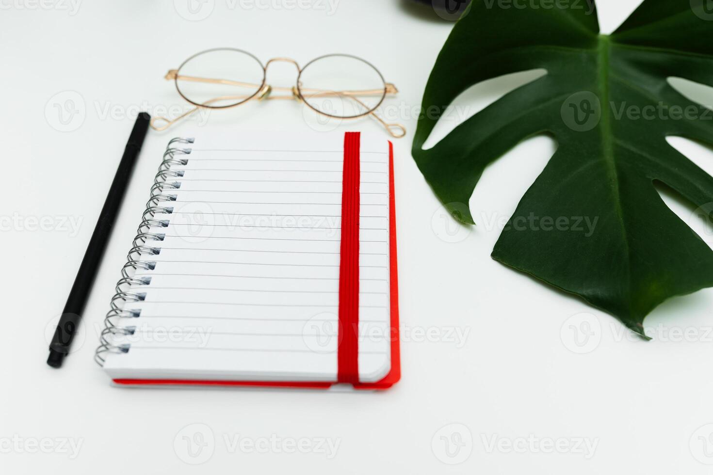 Open red notebook with pencil, glasses and monstera leaf. School notebook on white background, spiral notepad on table photo