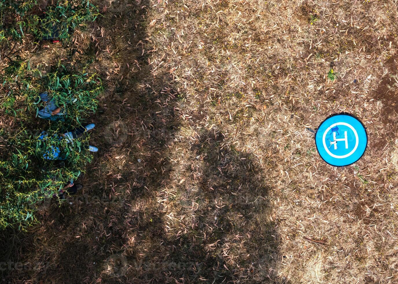 Drone landing pad surrounded by dried grass and scattered objects in a remote area as two persons sit underneath the shade photo