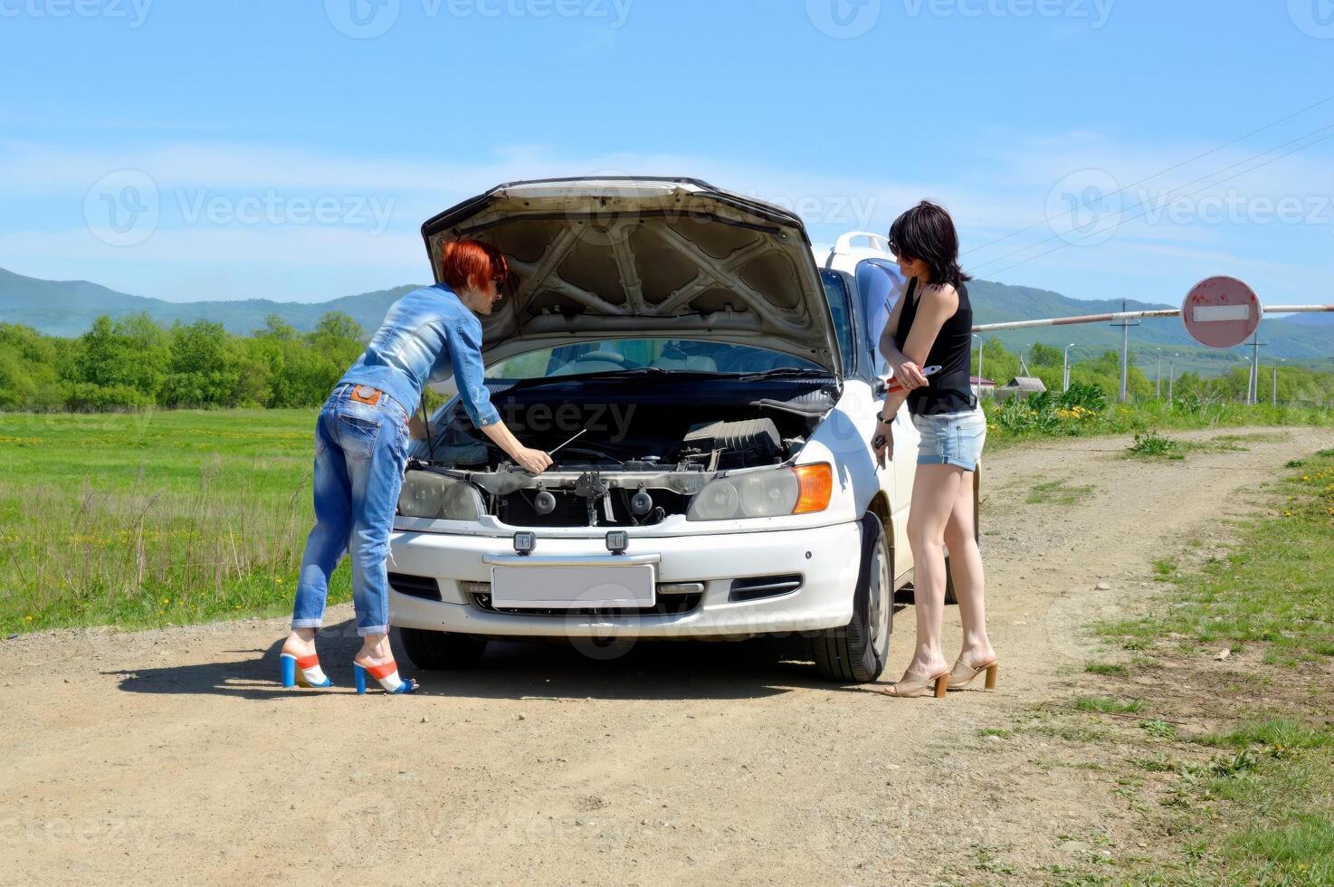 Red-haired woman with a screwdriver, on high heels standing near a broken car with open hood and brunette girl on a country road and barrier with stop sign photo