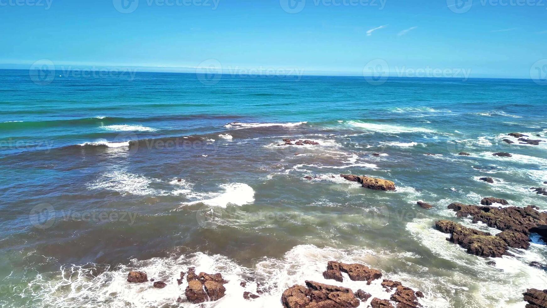 A stunning coastline where waves crash against rugged rocks under bright sunlight and clear blue skies photo