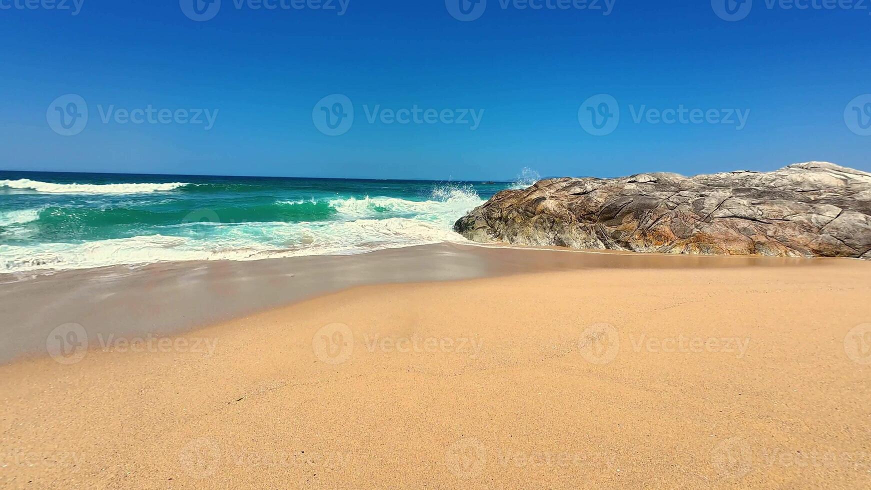 A picturesque beach scene featuring soft sand, gentle waves, and a large rock formation under a clear sky. photo