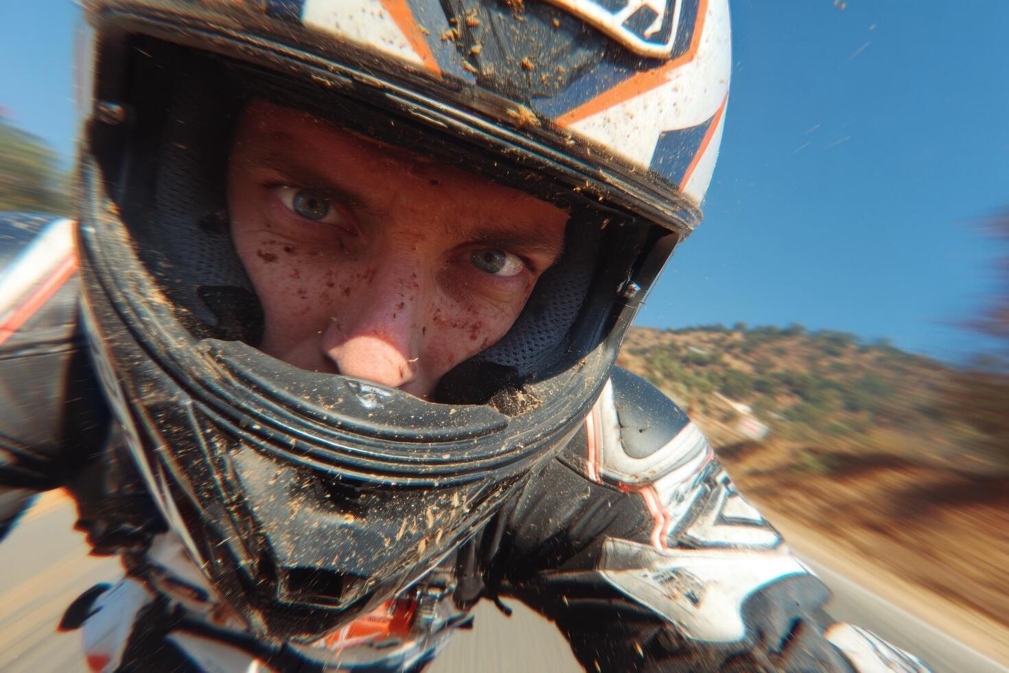 A motorcycle rider speeds along a dirt road, surrounded by mountains and clear blue skies, showcasing excitement and focus photo