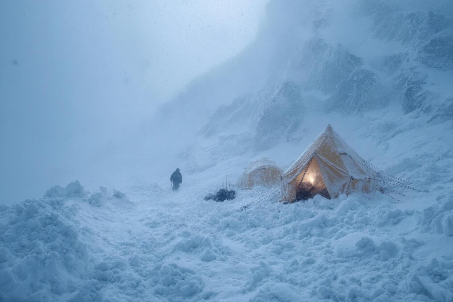 A person trudges through deep snow towards a tent amid heavy snowfall in a remote mountain location during winter photo