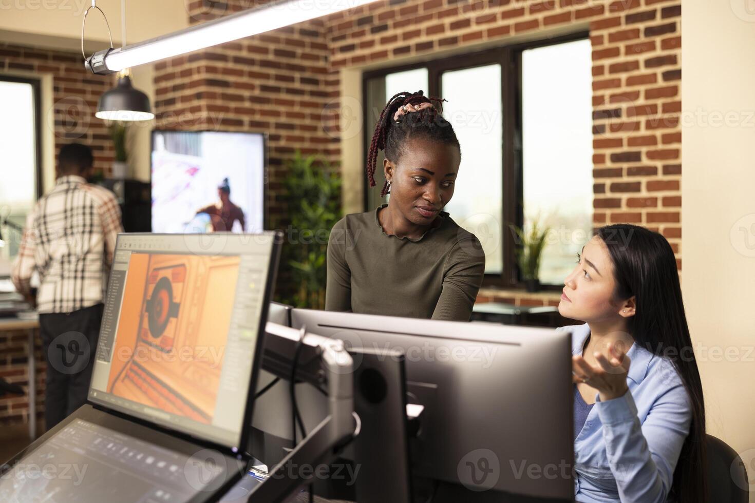 Professional women editors exchanging ideas, collaborating on a startup design project in brick wall office. Asian female employee sitting at desk and updating her black creative manager. photo