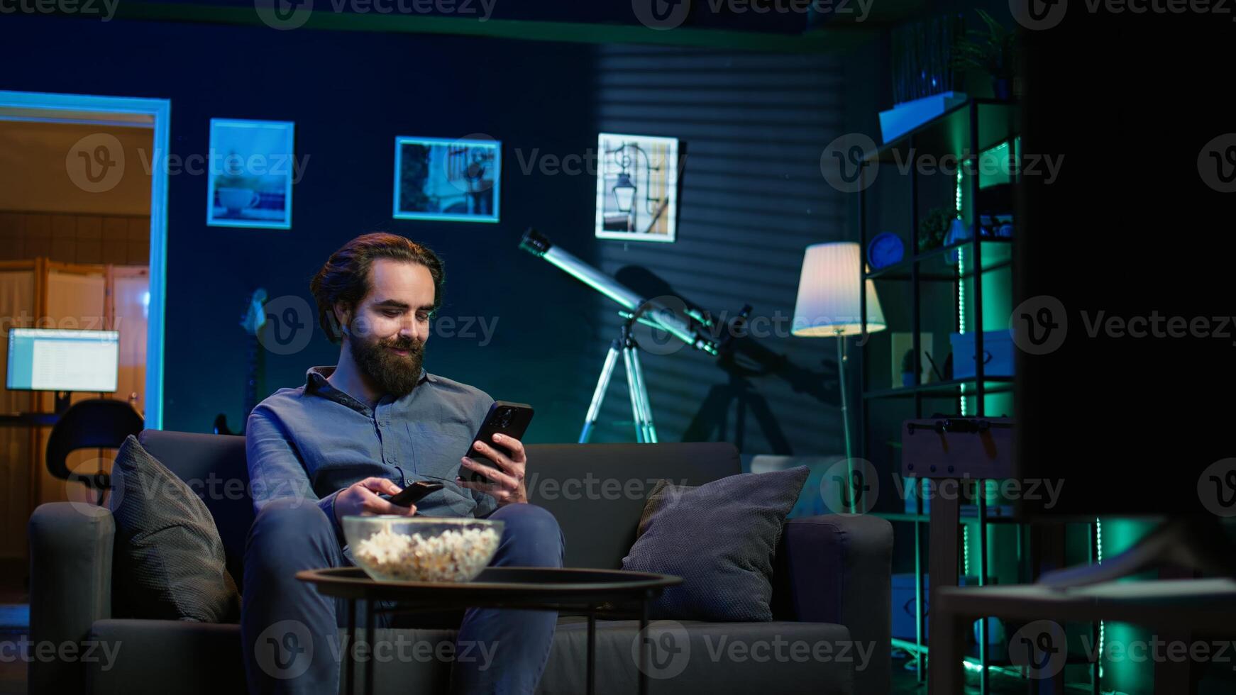 Smiling person on couch picking movie on streaming service using remote and texting on mobile phone. Happy man watching shows on television, zapping though channels, chatting on smartphone, camera B photo