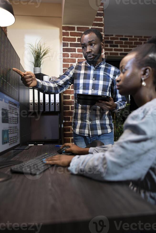 Male and female forex advisors working together at desk, analyzing stock market data on multiple screens late at night. The office features a brick wall and professional workstation. photo