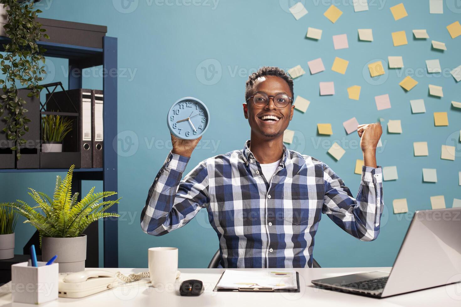 Happy businessman with a clock in hand, celebrating for finishing project presentation before deadline. Cheerful male manager raising fist with excitement, feeling great about end of workday. photo