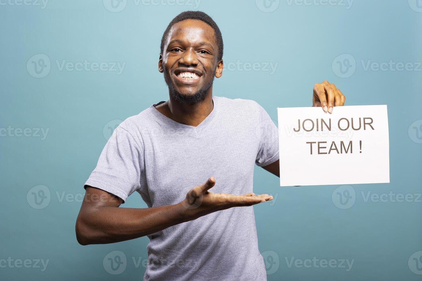 Enthusiastic recruiter looking at camera, holding a paper, inviting candidates to join his team. Smiling african american man pointing at hiring sign, promoting job openings in an isolated background. photo