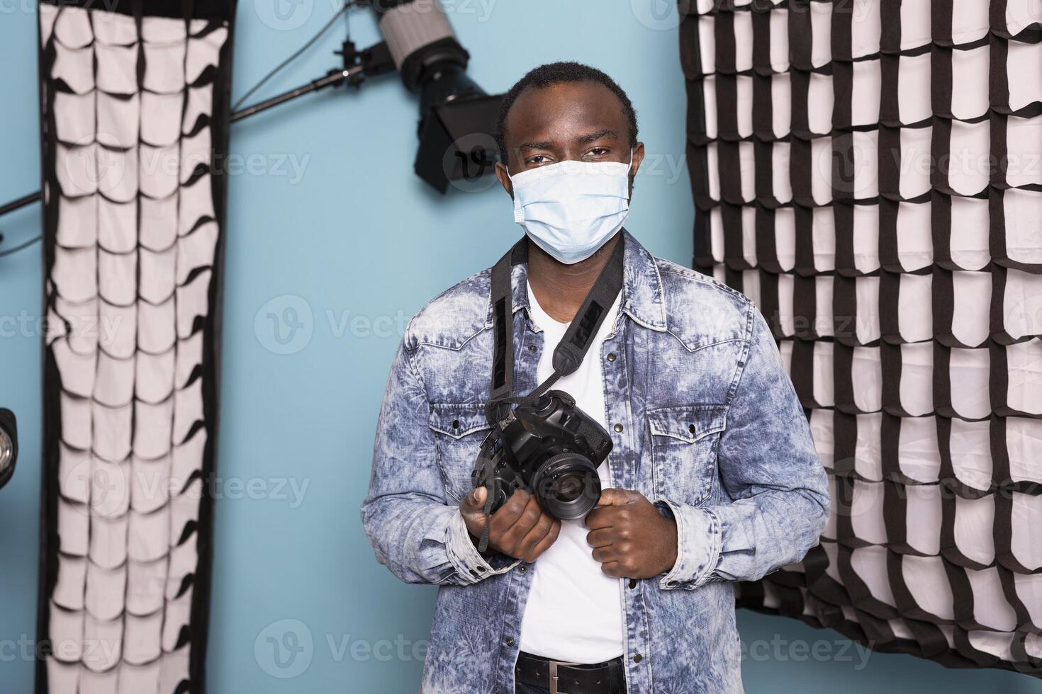 African american man holding his DSLR camera, wearing face mask as he prepares for creative project. Studio portrait of black photographer, taking precautions to protect himself from airborne diseases. photo